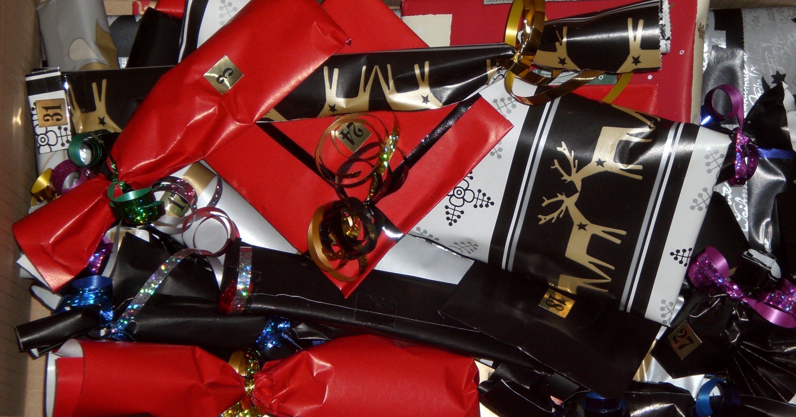 Karton voller einzeln verpackter Päckchen in Rot, Schwarz und Silber mit Geschenkband und kleinen goldenen Zahlenschildern, ein selbst befüllter Adventskalender