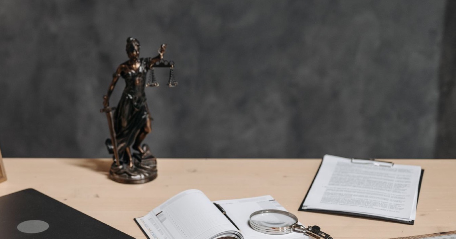 Justitia-Figur aus Bronze auf einem hellen Schreibtisch, daneben ein aufgeschlagener Terminkalender mit Lupe und ein Klemmbrett mit Dokumenten